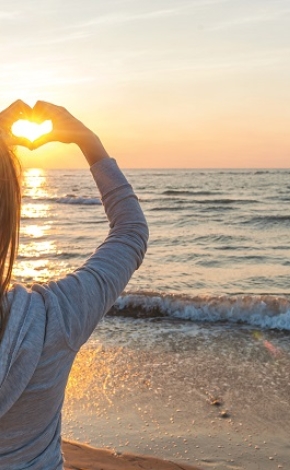 Girl standing on a beach making a hear shape with her hands facing the water