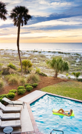 woman lounging on a pool floatie in a pool next to the beach during sunset